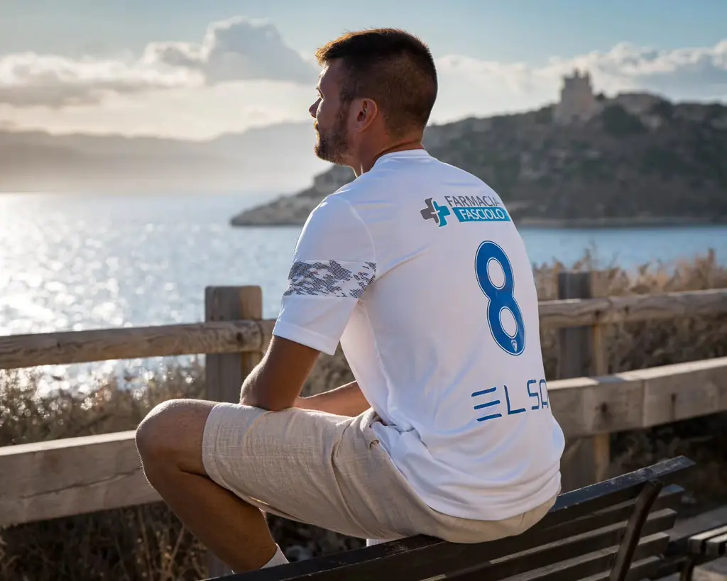 Un giocatore con la maglia bianca seduto davanti al mare e alla torre di Cagliari